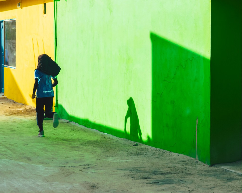 Girl running in The Maldives on the island of Thulusdoo against a green and yellow wall being chased by her shadow.
