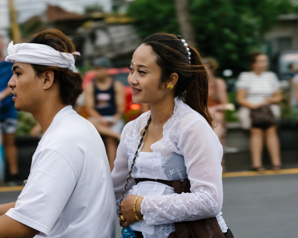 Balinese couple on a scooter during Ogoh Ogoh dressed in their finest clothes. The young woman is pregnant and holding her belly with one arm.