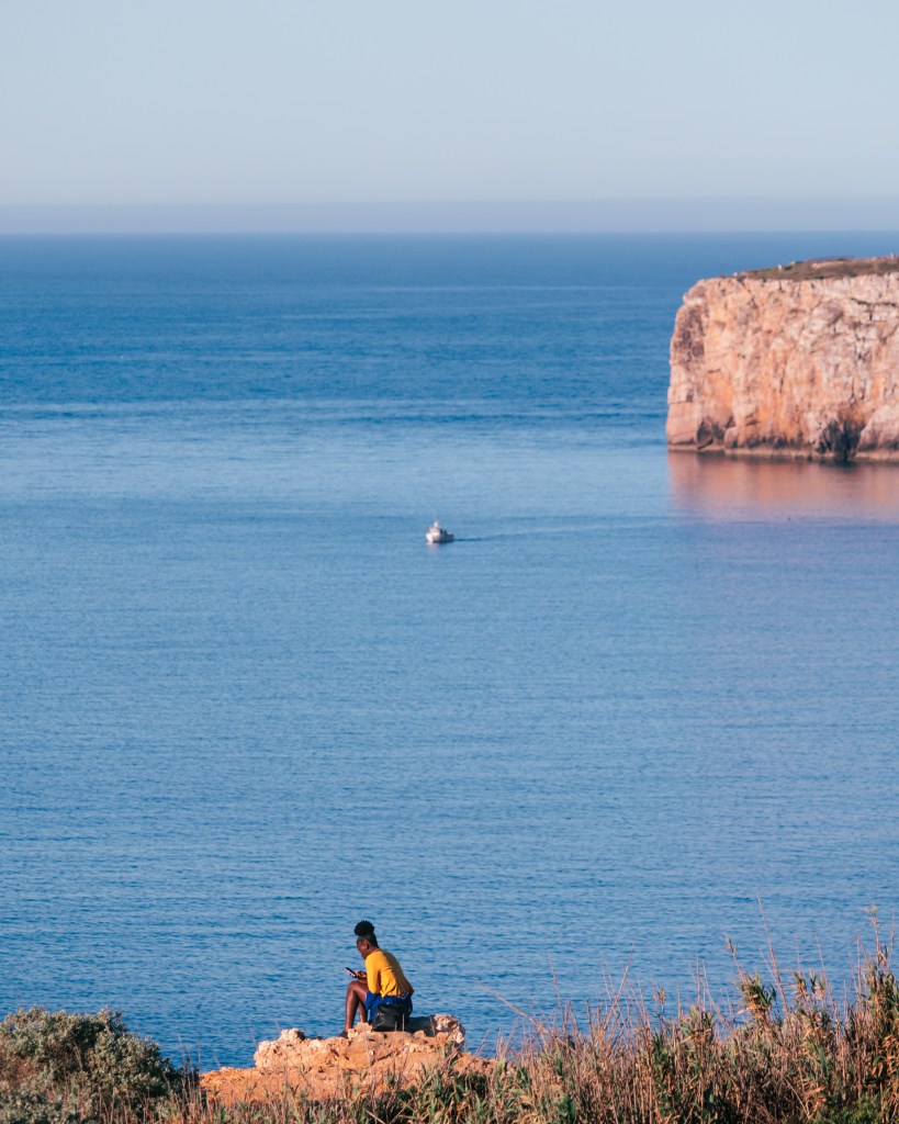 Woman sitting on a cliff in Sagres, Algarve, Portugal one april morning after sunrise checking how her selfies turned out in her phone