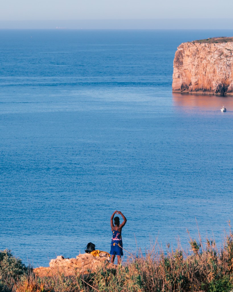 Woman standing on a cliff in Sagres, Algarve, Portugal one april morning after sunrise making a herat shape with her hands while taking a photo of it with her phone