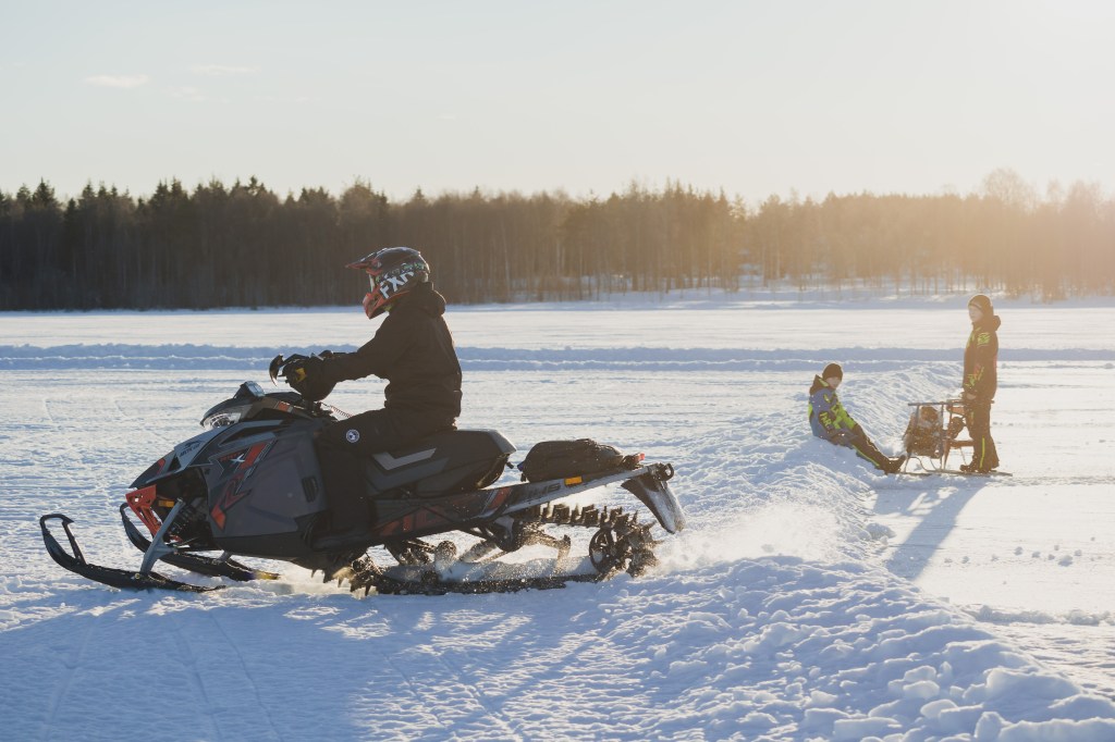 Young children on snow kick bikes watching a snow scooter drive by on Kalix River, Sweden, January 2023