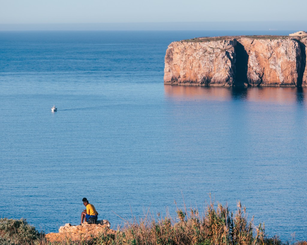Woman sitting on a cliff in Sagres, Algarve, Portugal one april morning after sunrise checking how her selfies turned out in her phone